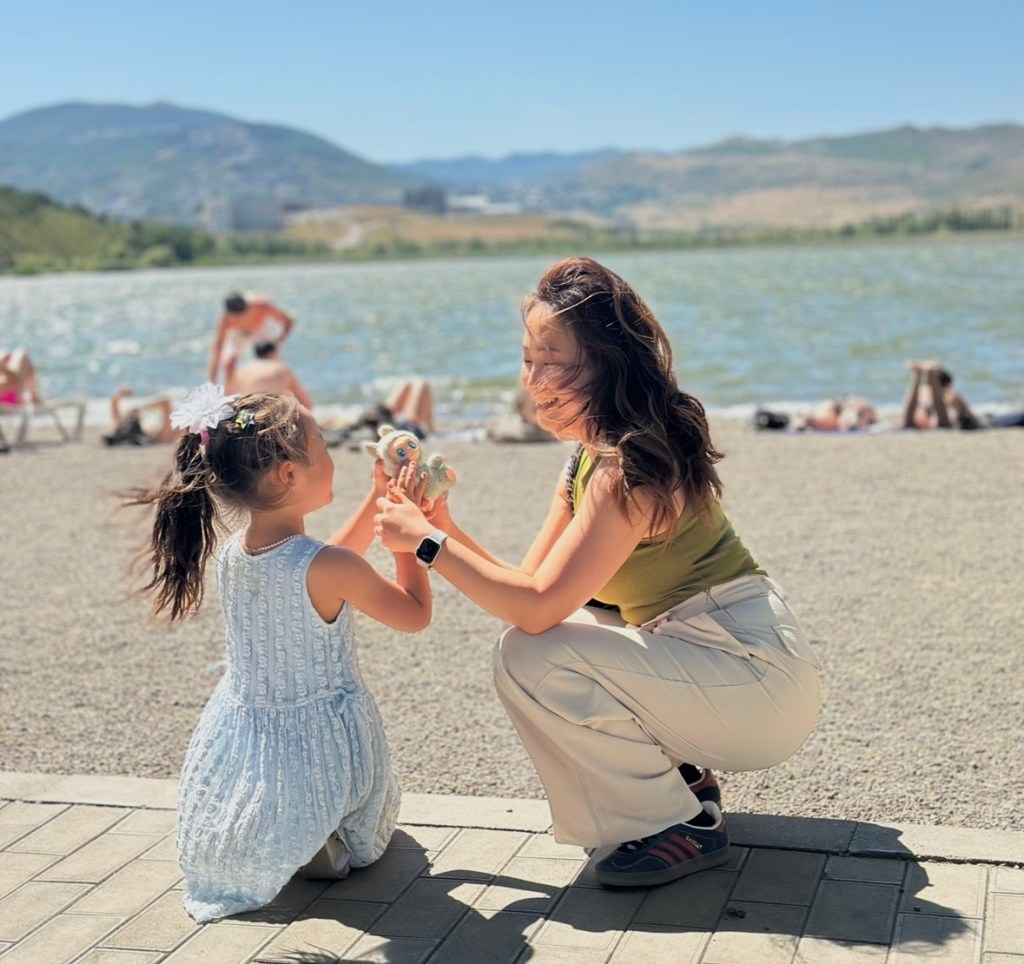 A woman and a young girl are playing by a lake on a sunny day. The girl, wearing a light blue dress, is kneeling and holding a toy, while the woman, in a green top and white pants, is crouching beside her, smiling.